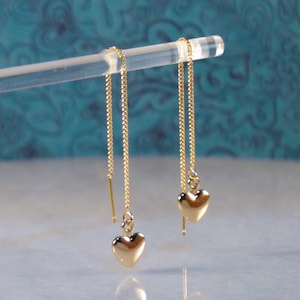 May include: A pair of gold heart-shaped dangle earrings with thin chains. The earrings are hanging from a clear acrylic display stand.