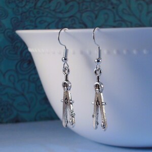 May include: Silver compass earrings with a delicate design. The earrings are hanging from a white surface.