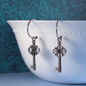 May include: A pair of silver-toned drop earrings featuring a key-shaped charm with an ornate, flower-like bow and a thin, curved wire hook for attachment. The metal has a darkened, antiqued finish that highlights the intricate details.