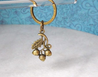 May include: A bronze-colored keychain featuring a decorative acorn and leaf design. The charm includes three acorns and a leaf, attached to a circular key ring. The keychain is suspended against a light blue patterned background.