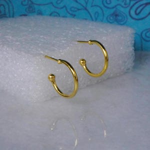 May include: A pair of small, gold-coloured hoop earrings. Each earring has a rounded, ball-shaped accent at the bottom and a straight post at the top. The earrings are displayed on a white surface.
