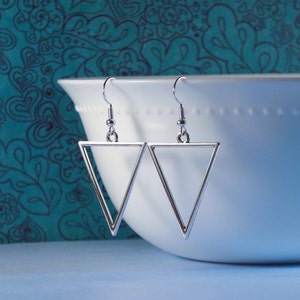 May include: A pair of silver triangle earrings with a simple geometric design. The earrings are hanging from a white bowl.