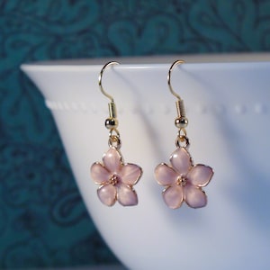 May include: A pair of gold dangle earrings with pink flower charms. The flowers have a white centre and are detailed with a light pink enamel.