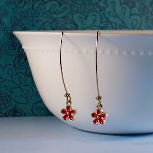 May include: A pair of gold dangle earrings with red flower charms. The earrings are hanging from a white bowl.