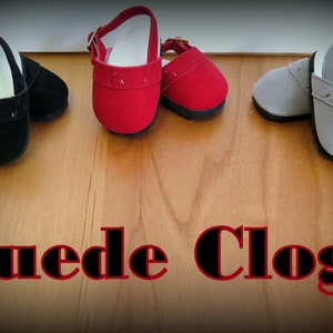 May include: Three pairs of suede doll shoes in black, red, and gray. The shoes have a strap and buckle closure. The shoes are on a wooden surface. "Suede Clogs" is written in black letters with a red outline below the shoes.