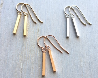 May include: Three pairs of minimalist bar earrings in gold, rose gold, and silver. Each pair features a simple, straight bar pendant hanging from a delicate hook.