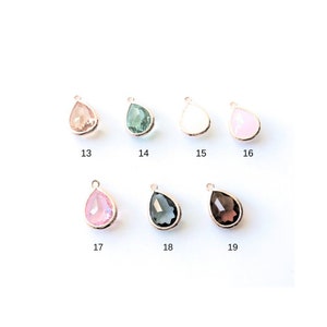 May include: Nine teardrop-shaped glass charms with gold-tone metal settings. The charms are in various colors, including pink, green, white, and gray.