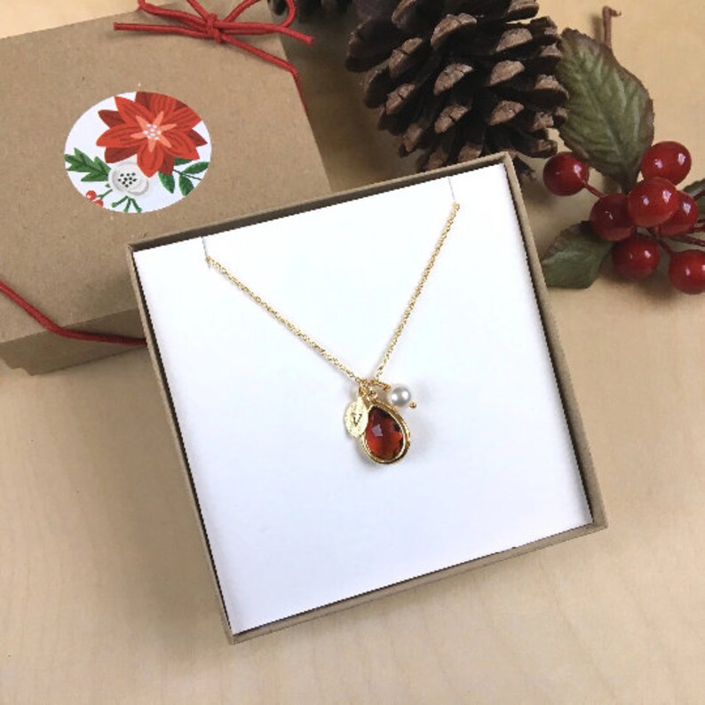 May include: A gold necklace with a teardrop-shaped red gemstone pendant, a small gold initial charm, and a pearl charm. The necklace is in a white gift box with a brown lid.