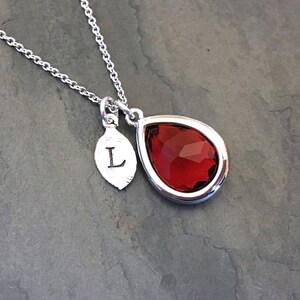 May include: A silver necklace with a red teardrop-shaped pendant and a small silver leaf charm with the letter "L" engraved on it.