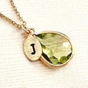 May include: A gold-tone necklace featuring a teardrop-shaped, faceted light green gemstone pendant paired with a small, oval-shaped gold-tone charm. The charm has a textured surface and displays the black capital letter J engraved on its front.