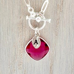 May include: Silver-toned necklace with a toggle clasp and a pendant. The pendant features a faceted, square-shaped, deep pink gemstone with a silver leaf charm and the letter "A". The necklace is displayed on a light wood surface.