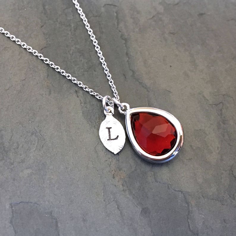 May include: A silver necklace with a teardrop-shaped pendant featuring a red gemstone. The pendant also has a small silver leaf charm with the letter "L" engraved on it.