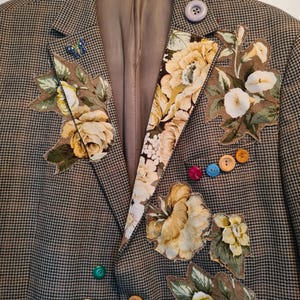 Könnte beinhalten: Ein braun-schwarzer Blazer mit Hahnentrittmuster und floralen Stoffapplikationen in Gelb, Creme und Grün. Der Blazer hat eine Vielzahl bunter Knöpfe und eine kleine Schmetterlingsnadel. Die Jacke hängt an einem schwarzen Kleiderbügel.