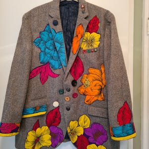 May include: A grey herringbone blazer featuring large, vibrant floral appliqués in shades of bright blue, orange, yellow, magenta, and purple. The garment is adorned with various colourful buttons scattered across the front and a small figurine pin.