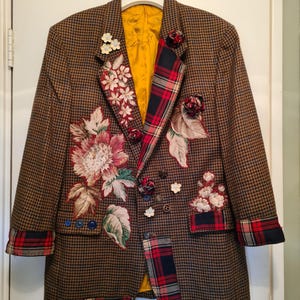 May include: A brown houndstooth blazer featuring a bright yellow lining, red and navy tartan fabric accents on the lapel, cuffs, and hem, and various floral fabric appliqués, small fabric rosettes, and decorative buttons across the front.