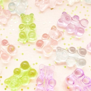 May include: Assortment of colorful gummy bear charms in various pastel shades, including pink, purple, green, and blue. The gummy bears are arranged on a light pink background with gold glitter.