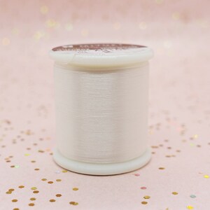 Könnte beinhalten: Eine Spule weißen Fadens in einem weißen Plastikbehälter. Der Behälter hat ein braunes Etikett mit dem Text "Extra Soft".