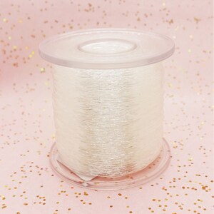 Könnte beinhalten: Eine Spule mit klarem, schimmerndem Garn, das auf einer Plastikspule aufgewickelt ist. Das Garn ist glänzend und hat einen dezenten Schimmer.