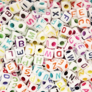 May include: A close-up of a pile of white plastic letter beads with colorful letters. The beads are all the same size and shape, and they are arranged in a random order.