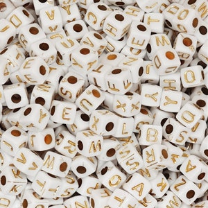 Può includere: Primo piano di perline quadrate bianche con lettere dorate. Ogni perlina mostra una lettera diversa dell'alfabeto. Le perline sono sparse e hanno fori per l'infilatura.