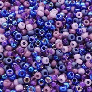 Może przedstawiać: Zbliżenie na kolekcję małych, okrągłych koralików w odcieniach niebieskiego, fioletowego i lawendowego. Koraliki mają otwór pośrodku, a niektóre mają metaliczny połysk. Koraliki są rozrzucone, tworząc teksturowaną powierzchnię.