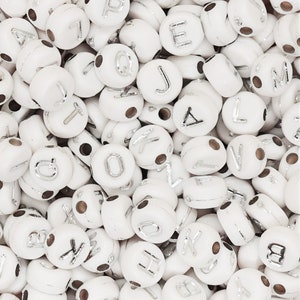 May include: A pile of white, spherical beads, each bearing a silver letter or number. The beads are scattered, displaying different letters. Each bead has a small hole for threading.