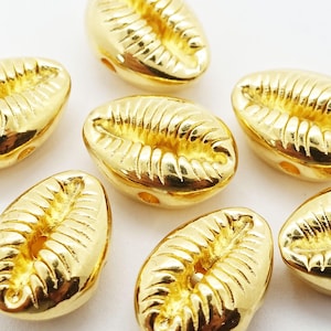 May include: Close-up of several gold-coloured cowrie shell beads. The beads have a glossy finish and are oval-shaped with detailed ridges. Each bead has two small holes for stringing. These beads are suitable for jewellery making and crafting.