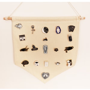 Könnte beinhalten: Eine weiße Leinwand-Tapete mit verschiedenen Emaille-Pins, die daran befestigt sind. Die Pins zeigen verschiedene Designs, darunter einen Schädel, eine Rose, einen Halbmond, ein Glas mit Sternen, eine Hand, einen Hexenhut, einen Sarg, eine Blume, eine Kaffeetasse, ein Herz und eine Katze.