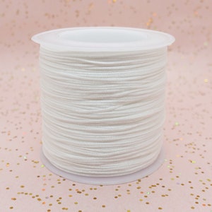 Pode incluir: Um carretel de cord&atilde;o branco, bem enrolado e apresentado sobre uma base de pl&aacute;stico branco. O cord&atilde;o parece liso e &eacute; adequado para v&aacute;rios projetos de artesanato. A imagem &eacute; um close-up, que destaca a textura e a cor do cord&atilde;o.