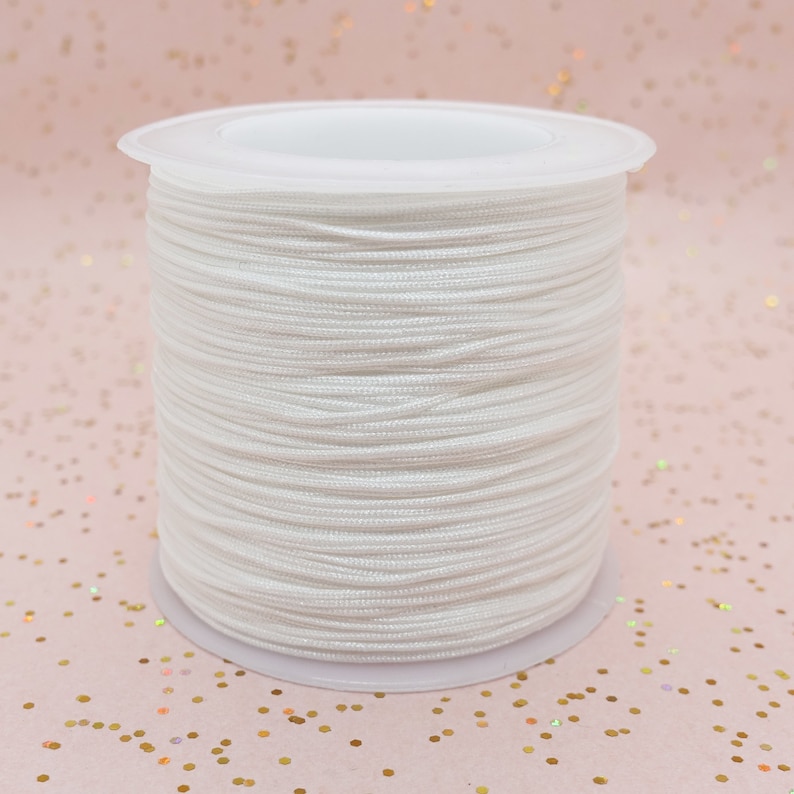 Pode incluir: Um carretel de cord&atilde;o branco, bem enrolado e apresentado sobre uma base de pl&aacute;stico branco. O cord&atilde;o parece liso e &eacute; adequado para v&aacute;rios projetos de artesanato. A imagem &eacute; um close-up, que destaca a textura e a cor do cord&atilde;o.