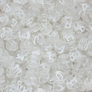 Pode incluir: Uma imagem de close-up de um monte de contas de plástico transparente com letras do alfabeto. As contas são redondas e têm letras brancas.
