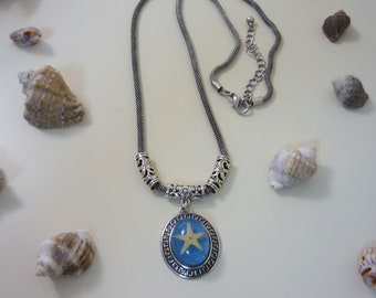 Puede incluir: Un collar de cadena plateada con un colgante azul y plateado con una estrella de mar dorada. El colgante está rodeado por un borde plateado con un diseño de llave griega.