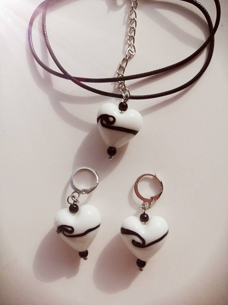 Puede incluir: Un collar de cord&oacute;n negro con un colgante de coraz&oacute;n blanco con un dise&ntilde;o de espiral negro. Pendientes a juego con colgantes de coraz&oacute;n blanco y dise&ntilde;os de espiral negro.