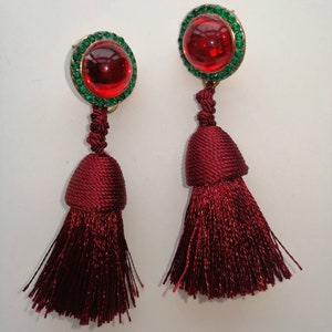 May include: A pair of red tassel earrings with a round, red glass cabochon set in a gold tone setting surrounded by green rhinestones.