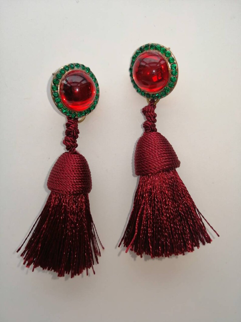 May include: A pair of red tassel earrings with a round, red glass cabochon set in a gold tone setting surrounded by green rhinestones.