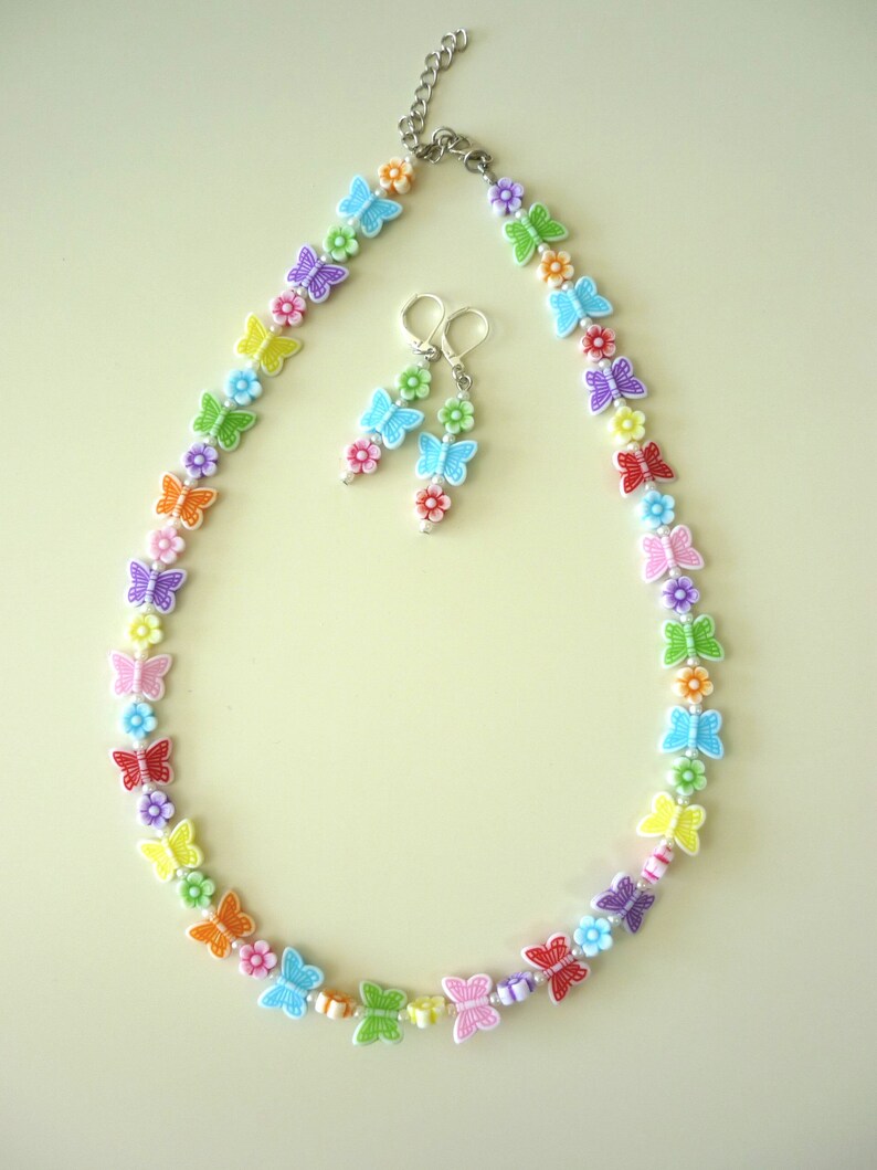 May include: A rainbow-colored necklace and earrings set featuring butterfly and flower charms. The necklace is made of plastic beads and has a silver chain closure. The earrings are dangle style with a butterfly charm at the bottom.