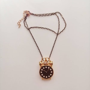 Puede incluir: Un collar dorado con un colgante de reloj negro. El reloj tiene una esfera negra con números y manecillas blancas, y está rodeado de pequeños diamantes de imitación. El collar tiene una cadena negra.
