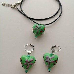 Puede incluir: Un collar con un colgante en forma de coraz&oacute;n verde y pendientes a juego. El colgante en forma de coraz&oacute;n y los pendientes tienen un dise&ntilde;o floral con detalles rojos, morados y turquesa.