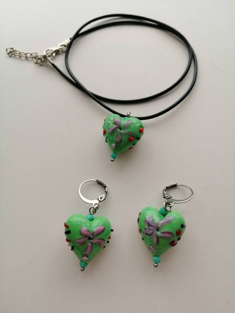 Puede incluir: Un collar con un colgante en forma de coraz&oacute;n verde y pendientes a juego. El colgante en forma de coraz&oacute;n y los pendientes tienen un dise&ntilde;o floral con detalles rojos, morados y turquesa.
