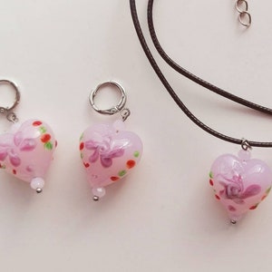 Puede incluir: Tres colgantes de coraz&oacute;n de vidrio rosa con dise&ntilde;os florales y lunares. Los colgantes est&aacute;n en un collar de cord&oacute;n negro y dos cadenas plateadas.