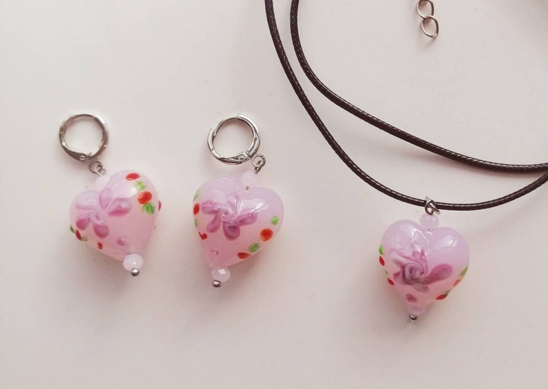 Puede incluir: Tres colgantes de coraz&oacute;n de vidrio rosa con dise&ntilde;os florales y lunares. Los colgantes est&aacute;n en un collar de cord&oacute;n negro y dos cadenas plateadas.