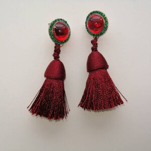 May include: A pair of red tassel earrings with green rhinestone accents. The earrings have a red cabochon stone in the center of the green rhinestones.