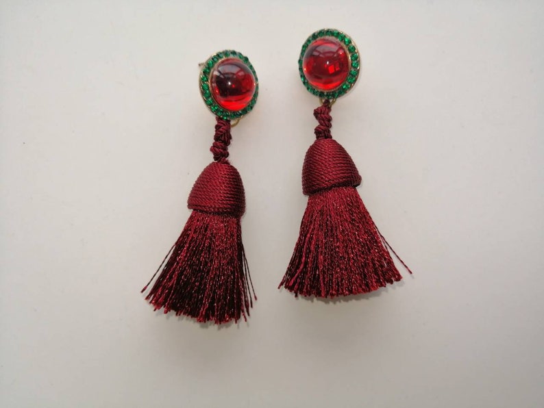 May include: A pair of red tassel earrings with green rhinestone accents. The earrings have a red cabochon stone in the center of the green rhinestones.