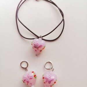 Puede incluir: Un conjunto de joyas de vidrio en forma de coraz&oacute;n rosa con dise&ntilde;os florales. El conjunto incluye un collar con un cord&oacute;n negro y un par de pendientes.