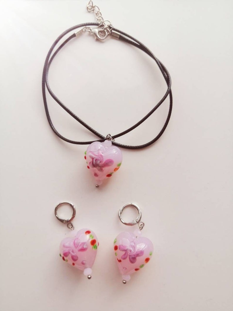 Puede incluir: Un conjunto de joyas de vidrio en forma de coraz&oacute;n rosa con dise&ntilde;os florales. El conjunto incluye un collar con un cord&oacute;n negro y un par de pendientes.