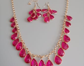 Puede incluir: Un conjunto de collar y pendientes a juego en tono dorado. El collar y los pendientes presentan colgantes vibrantes en forma de lágrima fucsia. Los colgantes están suspendidos de una delicada cadena dorada, creando un conjunto de joyas glamuroso.