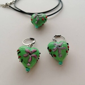 Puede incluir: Un collar con un colgante en forma de coraz&oacute;n verde y pendientes a juego con un dise&ntilde;o floral y detalles rojos y turquesa.