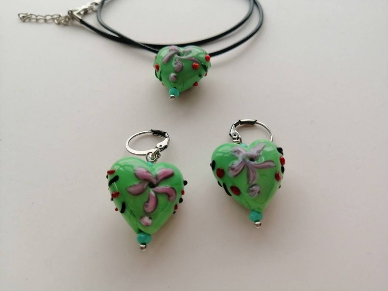 Puede incluir: Un collar con un colgante en forma de coraz&oacute;n verde y pendientes a juego con un dise&ntilde;o floral y detalles rojos y turquesa.