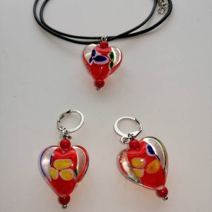 Puede incluir: Un conjunto de joyas de vidrio en forma de coraz&oacute;n rojo con un collar de cord&oacute;n negro y pendientes de aro de plata. Los corazones de vidrio tienen un fondo blanco con remolinos y dise&ntilde;os coloridos.