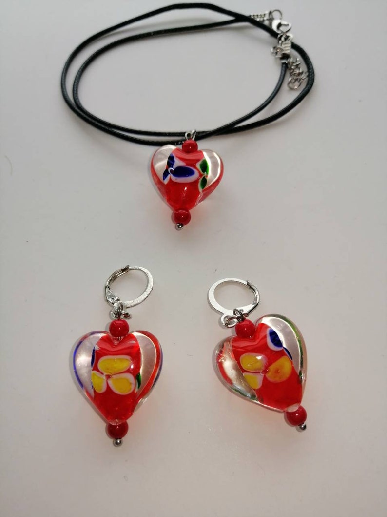 Puede incluir: Un conjunto de joyas de vidrio en forma de coraz&oacute;n rojo con un collar de cord&oacute;n negro y pendientes de aro de plata. Los corazones de vidrio tienen un fondo blanco con remolinos y dise&ntilde;os coloridos.